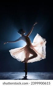 Theatrical performance. Beautiful, tender, graceful ballerina dancing against dark blue background with spotlight. Concept of art, classical ballet, creativity, choreography, beauty, ad