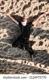 Theatrical Performance, Girl In White Theater Mask Lies On Sea Sand. Outdoor Theater Performance Of Woman Dancer At Sunny Day. Woman In White Mask And Total Black Style Of Cloth On Sea Coast, Top View