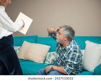 In their home's intimate setting, a senior couple grapples with distress as the woman gestures sternly towards documents, her frustration evident, while the man sits on the sofa - Powered by Shutterstock