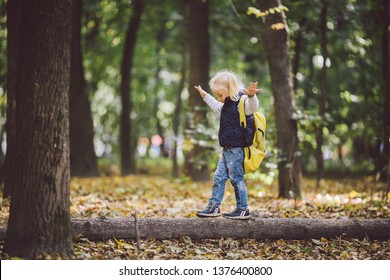 The Theme Children Outdoor Activities. Funny Little Baby Caucasian Blond Girl Walks Through Forest Overcoming Obstacles, Tree Fell, Log. Baby Hiking Big Funny Backpack In Autumn Forest Park.