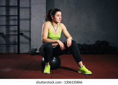 Theme Sport And Health. Strong Muscular Caucasian Woman In The Gym Sitting On A Black Heavy Padded Fitball Taking A Break From A Crossfit Strength Training In The Gym.