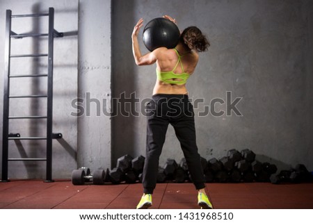 Theme sport and health. A strong muscular Caucasian woman in the gym trains strength and endurance. Equipping a large printed black ball raises and throws over her shoulder. Girl athlete crossfit.