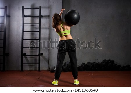 Theme sport and health. A strong muscular Caucasian woman in the gym trains strength and endurance. Equipping a large printed black ball raises and throws over her shoulder. Girl athlete crossfit.
