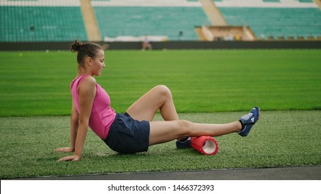 Theme Sport And Rehabilitation Sports Medicine. Teen Girl Athlete Uses A Massage Foam Roller Sitting On A Lawn Of A Football Field In A Stadium. Workout To Remove Pain, Stretch And Massage Muscles