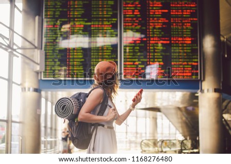 Theme travel and tranosport. Beautiful young caucasian woman in dress and backpack standing inside train station or terminal looking at a schedule holding a red phone, uses communication technology.