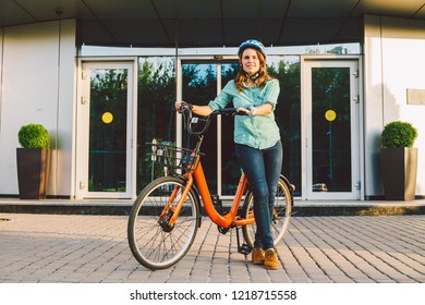 Theme to work on the bike. A young Caucasian woman arrived on environmentally friendly transport bike to the office. Girl in a bicycle parking office building in a helmet, gloves and shirt and jeans. - Powered by Shutterstock