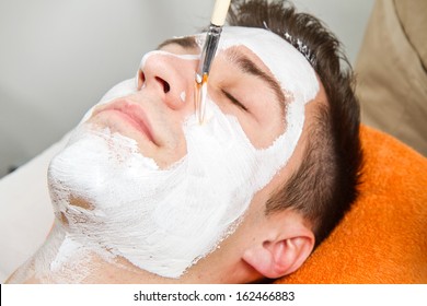Therapist applying a face mask to a beautiful young man in a spa using a cosmetics brush 