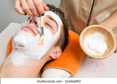 Therapist applying a face mask to a beautiful young man in a spa using a cosmetics brush 