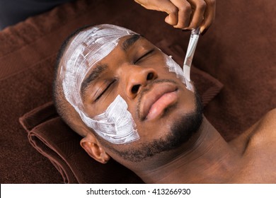 Therapist Applying Face Mask To Young African Man In Spa