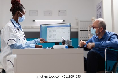 Therapist Doctor Wearing Protective Face Mask To Prevent Infection Cu Covid19 Discussing Disease Diagnosis With Senior Man In Wheelchair During Medical Appointment In Hospital Office. Medicine Concept