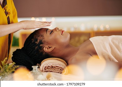 Therapist holding hands over head of pretty young Black woman to transfer energy during healing reiki session