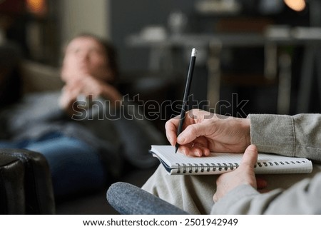 Therapist holding pen and notepad while patient sits in background with blurred face. Focus is on therapist's hand and pen without showing patient's details