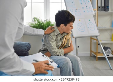 Therapist trying to help unhappy, resentful kid who has behaviour issues. Portrait of sad caucasian boy hugging wooden man figurine to relieve stress in psychologists office. Psychotherapist interior. - Powered by Shutterstock