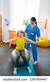 Therapist In Uniform. Hard-working Long-haired Girl In Yellow T-shirt Stretching Rope Between Her Hands While Saving Balance On Pilates Ball