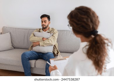 In a therapy session, middle eastern man seeking help clutches a pillow while talking to psychologist, conveying vulnerability