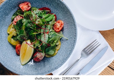 There Are A Blue Deep Bowl Of Salad On The Table Of The Vegetarian Restaurant. It Looks Very Healthy An Wonderful. The Dish Is Served To People. In The Bowl There Are A Lot Of Leafs And Vegetables.