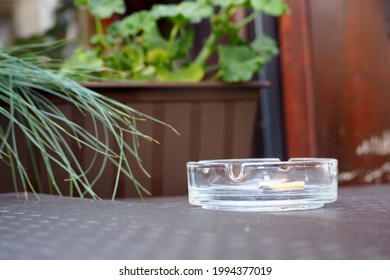 There Is A Cigarette Butt In A Round Glass Ashtray. Front View. There Is An Ashtray On A Dark Metal Table In A Cafe