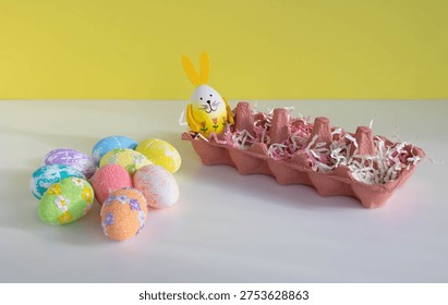 There is a container on the table, and Easter eggs of different colors are lying next to it, including one with bunny ears