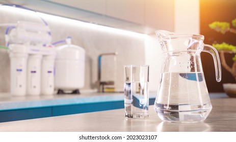 There Is A Decanter And A Glass Of Clean Water On The Kitchen Table. In The Background Water Filtration System