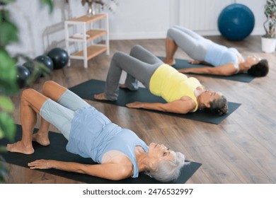 There is group Pilates class in studio for people of different ages. Elderly women in company of hatha yoga lovers performs bridge pose, setu bandha sarvangasana - Powered by Shutterstock