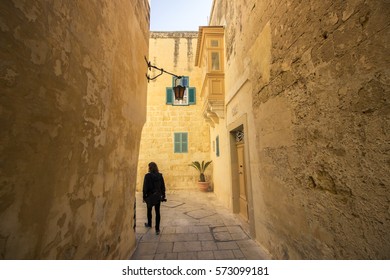 There is a guy on the image from behind, walking a narrow street between ancient Maltese buildings. 