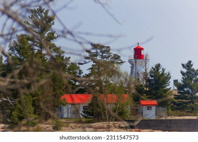 There is a lighthouse in the back of the picture in front of the picture there are some trees the lighthouse is white and it has a red roof there is a little cabin beside the lighthouse - Shutterstock ID 2311547573
