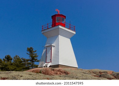 There is a lighthouse at the top of a hill there are some trees around it the lighthouse is with and it has a red roof the door is white the sky is blue on a sunny day - Shutterstock ID 2311547595
