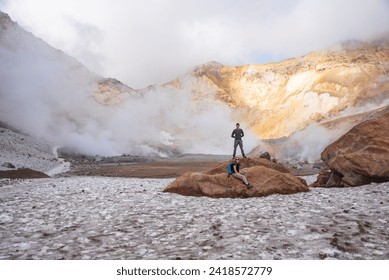 There are man and woman on the foreground. It is Mutnovskiy voclano. There is a field of fumaroles on the background.