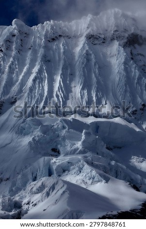 There are mountains that invite, and there are mountains that warn,  and the south face of Chacraraju, captured here in a brooding, steel-blue study of glacial architecture at its most merciless and magnificent, belongs unambiguously, irrevocably to the second category. Emerging from a shroud of fast-moving Andean cloud like a fortress wall built by forces indifferent to human existence, the upper ramparts of this 6,112-meter Cordillera Blanca giant reveal themselves in this extraordinary image as a vertical world of compressed seracs, hanging ice mushrooms, razor-edged snow flutings and fractured glacier blocks balanced in states of geological tension that could resolve, violently, instantaneously and without warning,  at any moment. The south face is not a climb for the ambitious or the brave, it is a climb for the rare, committed, technically masterful alpinist who has already proven everything there is to prove on lesser objectives and arrives here with eyes completely open to the scale of consequence that this wall demands as its entry fee. Every vertical channel of ice-carved snow fluting visible across the upper face represents a natural avalanche funnel; every hanging serac in the chaotic icefall below the headwall is a multi-ton projectile in a state of permanent, slow-motion gravitational negotiation. The cold, flat light of an approaching storm drains the scene of warmth and contrast, reducing the mountain to an almost monochromatic study in blue-grey ice, shadow and raw, architectural power, a palette that feels less like weather and more like the mountain itself issuing a final, unambiguous assessment of the situation to anyone below with the experience to read it correctly. For alpinists, adventure photographers, extreme climbing documentarians and anyone who understands the peculiar, addictive and deeply human compulsion to stand beneath something genuinely terrifying and choose to continue upward anyway