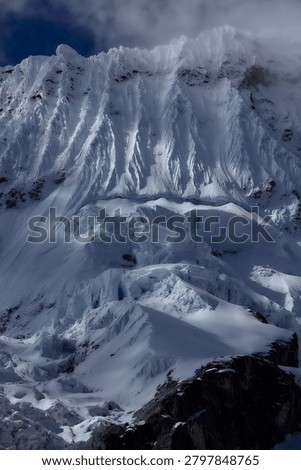 There are mountains that invite, and there are mountains that warn,  and the south face of Chacraraju, captured here in a brooding, steel-blue study of glacial architecture at its most merciless and magnificent, belongs unambiguously, irrevocably to the second category. Emerging from a shroud of fast-moving Andean cloud like a fortress wall built by forces indifferent to human existence, the upper ramparts of this 6,112-meter Cordillera Blanca giant reveal themselves in this extraordinary image as a vertical world of compressed seracs, hanging ice mushrooms, razor-edged snow flutings and fractured glacier blocks balanced in states of geological tension that could resolve, violently, instantaneously and without warning,  at any moment. The south face is not a climb for the ambitious or the brave, it is a climb for the rare, committed, technically masterful alpinist who has already proven everything there is to prove on lesser objectives and arrives here with eyes completely open to the scale of consequence that this wall demands as its entry fee. Every vertical channel of ice-carved snow fluting visible across the upper face represents a natural avalanche funnel; every hanging serac in the chaotic icefall below the headwall is a multi-ton projectile in a state of permanent, slow-motion gravitational negotiation. The cold, flat light of an approaching storm drains the scene of warmth and contrast, reducing the mountain to an almost monochromatic study in blue-grey ice, shadow and raw, architectural power, a palette that feels less like weather and more like the mountain itself issuing a final, unambiguous assessment of the situation to anyone below with the experience to read it correctly. For alpinists, adventure photographers, extreme climbing documentarians and anyone who understands the peculiar, addictive and deeply human compulsion to stand beneath something genuinely terrifying and choose to continue upward anyway