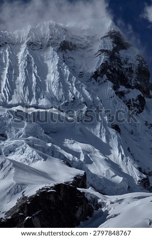 There are mountains that invite, and there are mountains that warn,  and the south face of Chacraraju, captured here in a brooding, steel-blue study of glacial architecture at its most merciless and magnificent, belongs unambiguously, irrevocably to the second category. Emerging from a shroud of fast-moving Andean cloud like a fortress wall built by forces indifferent to human existence, the upper ramparts of this 6,112-meter Cordillera Blanca giant reveal themselves in this extraordinary image as a vertical world of compressed seracs, hanging ice mushrooms, razor-edged snow flutings and fractured glacier blocks balanced in states of geological tension that could resolve, violently, instantaneously and without warning,  at any moment. The south face is not a climb for the ambitious or the brave, it is a climb for the rare, committed, technically masterful alpinist who has already proven everything there is to prove on lesser objectives and arrives here with eyes completely open to the scale of consequence that this wall demands as its entry fee. Every vertical channel of ice-carved snow fluting visible across the upper face represents a natural avalanche funnel; every hanging serac in the chaotic icefall below the headwall is a multi-ton projectile in a state of permanent, slow-motion gravitational negotiation. The cold, flat light of an approaching storm drains the scene of warmth and contrast, reducing the mountain to an almost monochromatic study in blue-grey ice, shadow and raw, architectural power, a palette that feels less like weather and more like the mountain itself issuing a final, unambiguous assessment of the situation to anyone below with the experience to read it correctly. For alpinists, adventure photographers, extreme climbing documentarians and anyone who understands the peculiar, addictive and deeply human compulsion to stand beneath something genuinely terrifying and choose to continue upward anyway