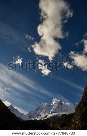 There are mountains that invite, and there are mountains that warn,  and the south face of Chacraraju, captured here in a brooding, steel-blue study of glacial architecture at its most merciless and magnificent, belongs unambiguously, irrevocably to the second category. Emerging from a shroud of fast-moving Andean cloud like a fortress wall built by forces indifferent to human existence, the upper ramparts of this 6,112-meter Cordillera Blanca giant reveal themselves in this extraordinary image as a vertical world of compressed seracs, hanging ice mushrooms, razor-edged snow flutings and fractured glacier blocks balanced in states of geological tension that could resolve, violently, instantaneously and without warning,  at any moment. The south face is not a climb for the ambitious or the brave, it is a climb for the rare, committed, technically masterful alpinist who has already proven everything there is to prove on lesser objectives and arrives here with eyes completely open to the scale of consequence that this wall demands as its entry fee. Every vertical channel of ice-carved snow fluting visible across the upper face represents a natural avalanche funnel; every hanging serac in the chaotic icefall below the headwall is a multi-ton projectile in a state of permanent, slow-motion gravitational negotiation. The cold, flat light of an approaching storm drains the scene of warmth and contrast, reducing the mountain to an almost monochromatic study in blue-grey ice, shadow and raw, architectural power, a palette that feels less like weather and more like the mountain itself issuing a final, unambiguous assessment of the situation to anyone below with the experience to read it correctly. For alpinists, adventure photographers, extreme climbing documentarians and anyone who understands the peculiar, addictive and deeply human compulsion to stand beneath something genuinely terrifying and choose to continue upward anyway