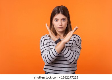 There Is No Way. Portrait Of Determined Serious Woman With Brown Hair In Long Sleeve Striped Shirt Standing With Crossed Arms And Gesturing No Sign. Indoor Studio Shot Isolated On Orange Background