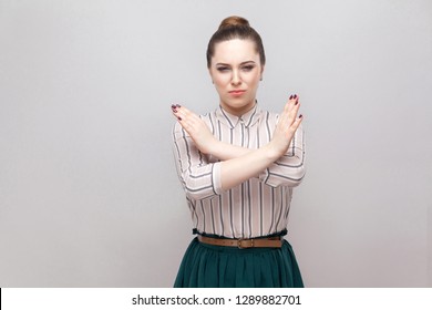 There is no way. Portrait of serious beautiful young woman in striped shirt and green skirt and collected ban hairstyle, standing with block, X sign. indoor studio shot, isolated on grey background.