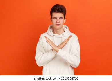 There is no way! Serious concentrated young man in stylish hoodie showing no gesture crossing arms, say no to bad habits, addiction. Indoor studio shot isolated on orange background