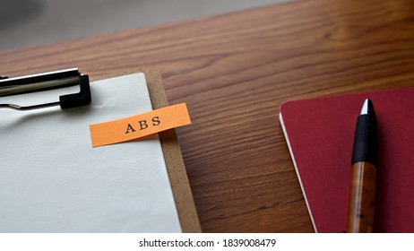 There Is A Notebook, A Pen, And A Clipborad With A Sticky Note Stuck To It That Says ABS Written On It. It Was An Abbreviation For Asset Backed Security.