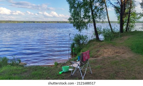 There Is A Place For Fishing On The Lake Shore. There Is A Folding Chair, A Stand For A Feeder Rod, A Bucket With Bait And A Box With Baits. A Towel Is Hanging On The Back Of The Chair.