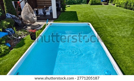 There is a rectangular frame PVC swimming pool with clean water on the mown lawn. Nearby are flower beds, bushes, thujas, swing, wooden arbor with table and benches. Sunny summer weather