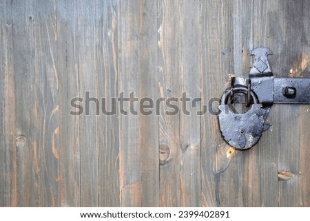 There is a rusty lock hanging on the wooden gate. Close-up view of a closed door. The wooden door is locked. A crude lock on the door, painted black.
