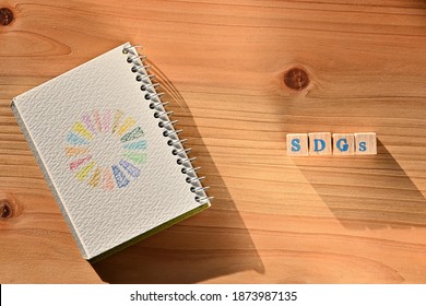 There is sketchbook with the illustration of Symbol of SDGs on it and word cube beside formed SDGs.