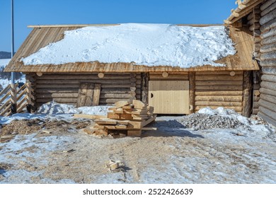 There is snow on the roof of the house. Renovated rustic house built in the 19th century. Cracked dark logs of a wooden house. Flat boards near the house in the countryside. - Powered by Shutterstock