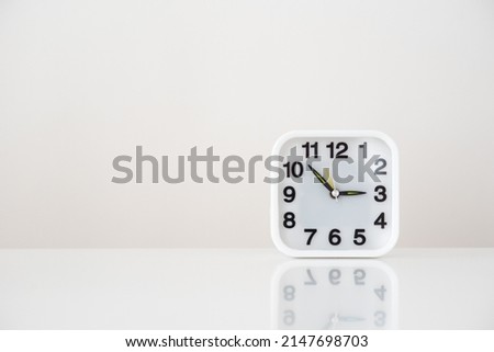 there is a square white clock with black numbers and arrows on a white table on a white background, free space for insertion, a mirror image on the table