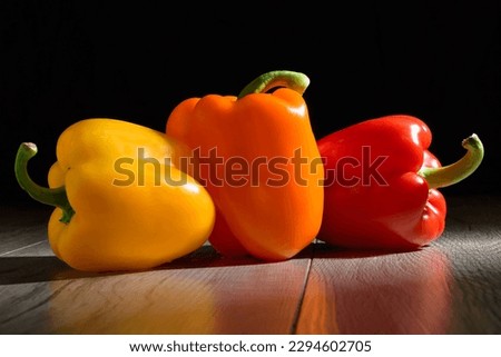 There are three colorful bell peppers on the wooden table. There is  dark photography of  yellow, red and orange capsicums.