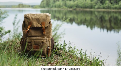 There Is A Tourist's Brown Backpack On The Shore Of A River. A Great Trip To Nature In A Dense Green Forest With A Reservoir. A Quiet And Picturesque Place For Reflection And Relaxation.