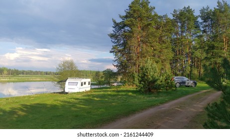 There are trees on the grass-covered lawn on the river bank, and a caravan trailer stands for convenience while fishing. A crossover vehicle stands by the dirt road. On the opposite bank is a forest