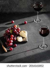 There Are Two Glasses Of Wine, Cheese And Grapes On The Dark Table, The Sun Is Shining Outside The Window. French Cheese And Red Wine On A Gray Background. Still Life With Alcohol And Fruit