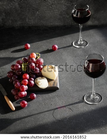 There are two glasses of wine, cheese and grapes on the dark table, the sun is shining outside the window. French cheese and red wine on a gray background. Still life with alcohol and fruit