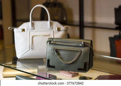 There Are Two Handbags On A Glass Shelf In The Store. One Bag Is Green Or Khaki, The Second Bag Is White. They Are Made Of Leather.