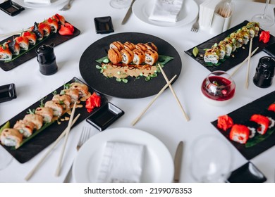 There Is A White Table With A Lot Of Plates With Sushi On It In The Restaurant. They All Look Very Delicious And Yummy. The Table Is For A Lot Of People. We Can See A Lot Of Different Types Of Sushi.