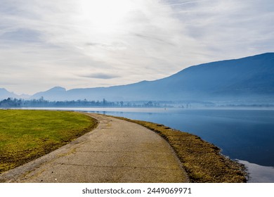 There is a wonderful mountain, lake, forest, fog reflection image in the background, and a walking path and green field in a bright light in the foreground - Powered by Shutterstock